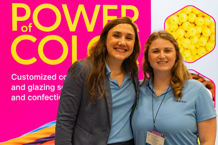 two women in light blue shirts standing in front of a bright pink sign at their booth